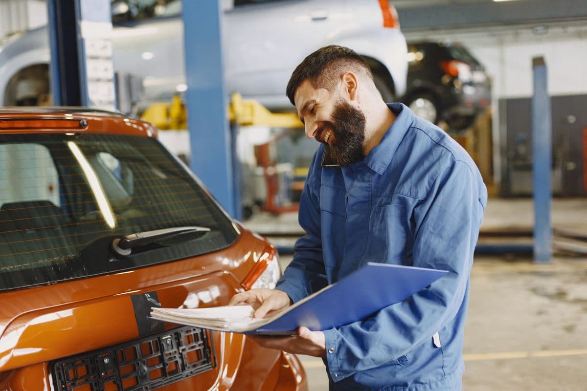 Mechaniker prüft ein Fahrzeug in der Werkstatt und hält die Unterlagen dazu in der Hand